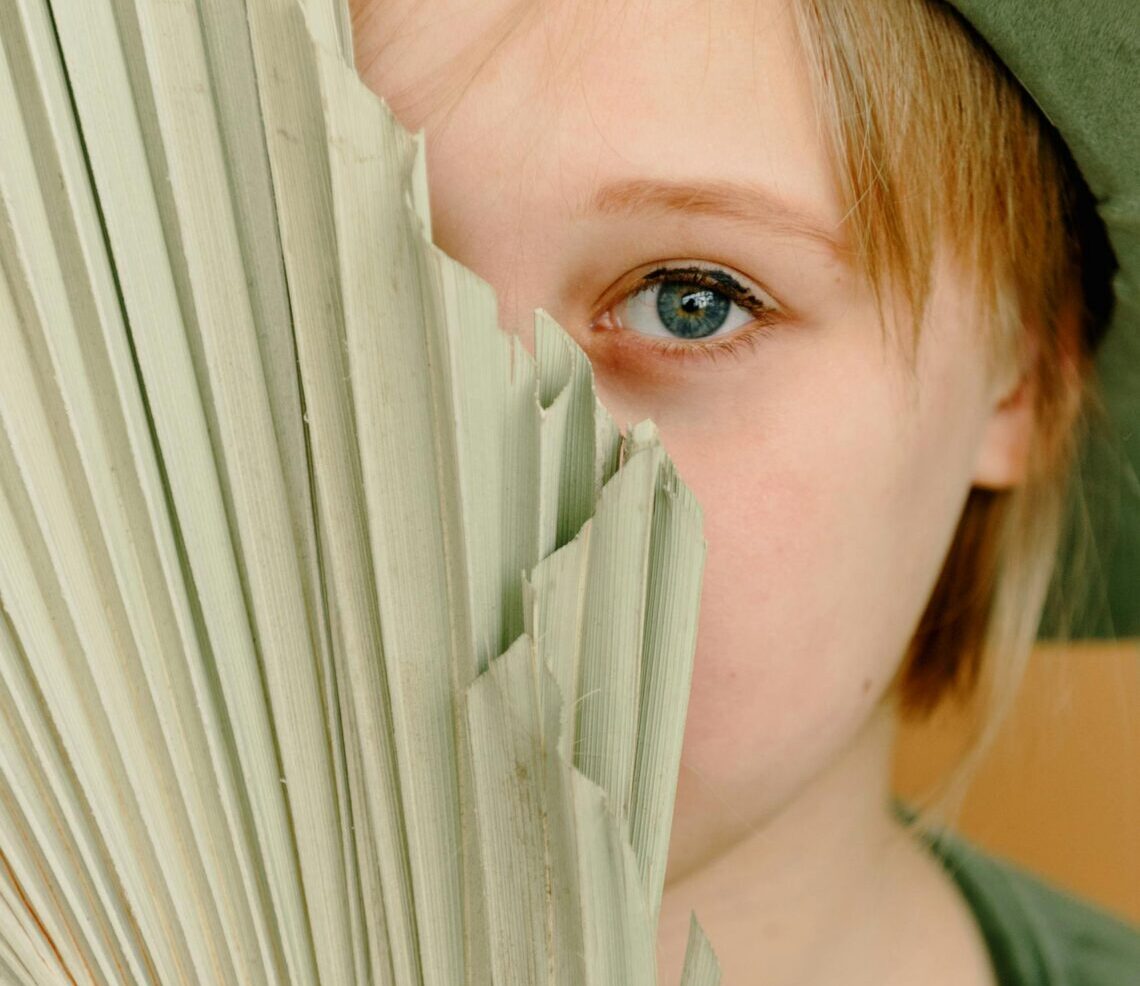 A child playfully hides behind a dried palm leaf, capturing an innocent and curious expression.