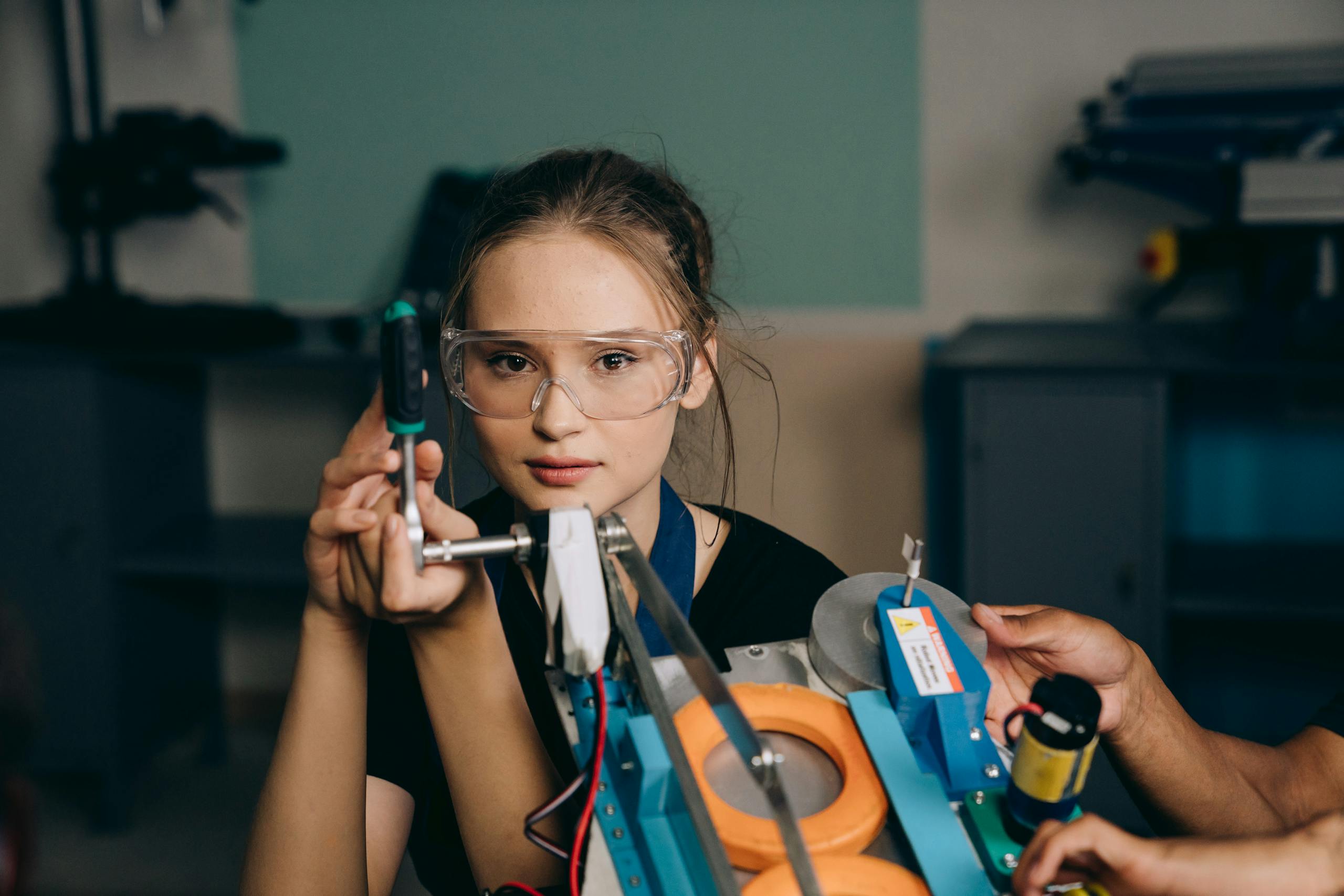 A young woman with goggles uses a tool in a workshop environment, focusing intently on her task.