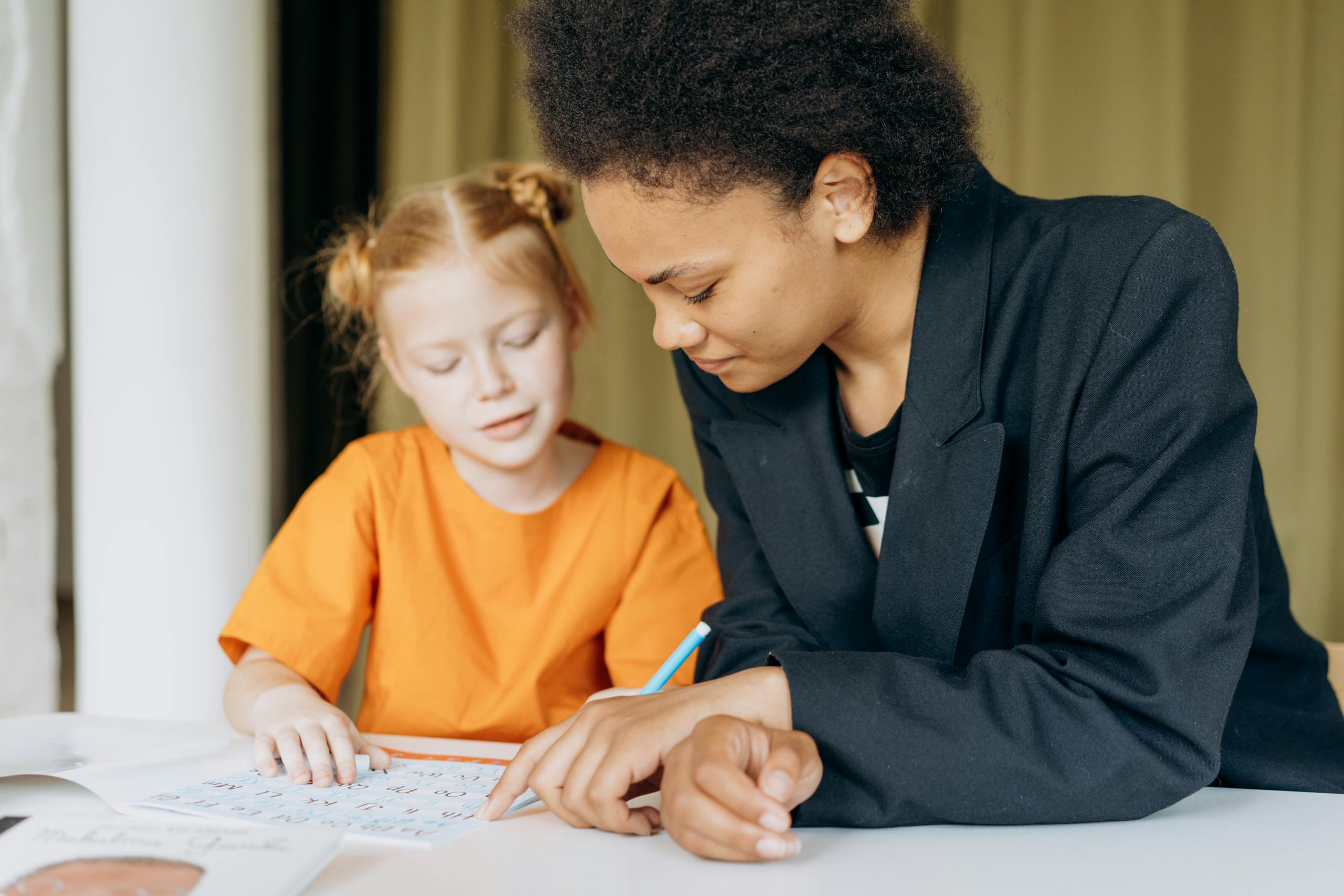 A teacher helps a young student with writing tasks at a desk indoors.