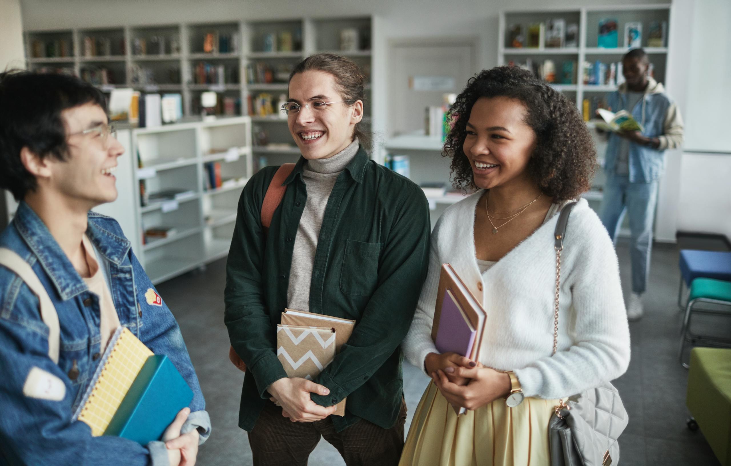 Photo by Andy Barbour Group of diverse students laughing and holding books in a library environment.